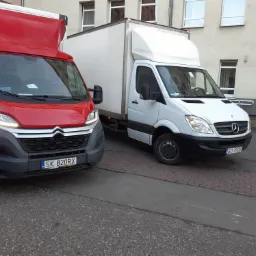 Dwa busy dostawcze: czerwony Citroen i biały Mercedes, zaparkowane na szarym chodniku przed budynkiem.