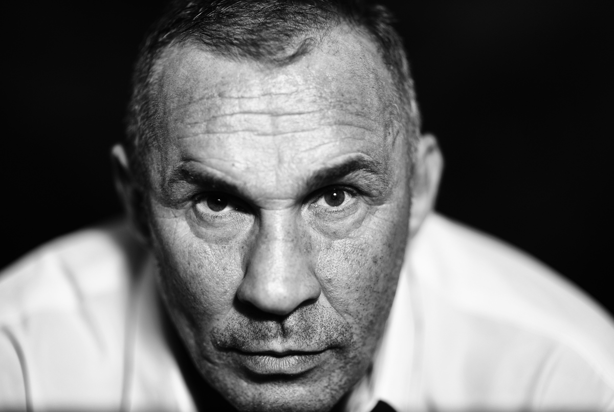 Czarno-biały portret mężczyzny z bliska, ostre światło podkreśla zmarszczki i rysy twarzy, intensywne spojrzenie w obiektyw.