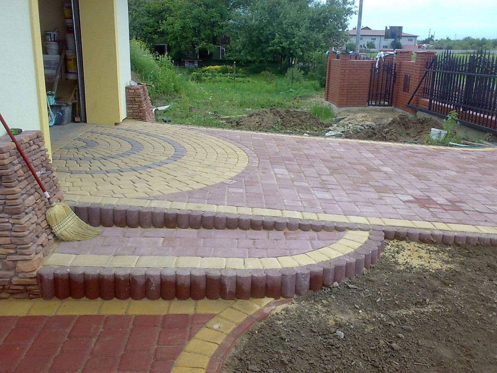 Ułożona kostka brukowa w odcieniach czerwieni i beżu tworząca wzór na podjeździe z niskimi schodami, widoczna miotła oparta o ścianę budynku, w tle trawnik i ogrodzenie.