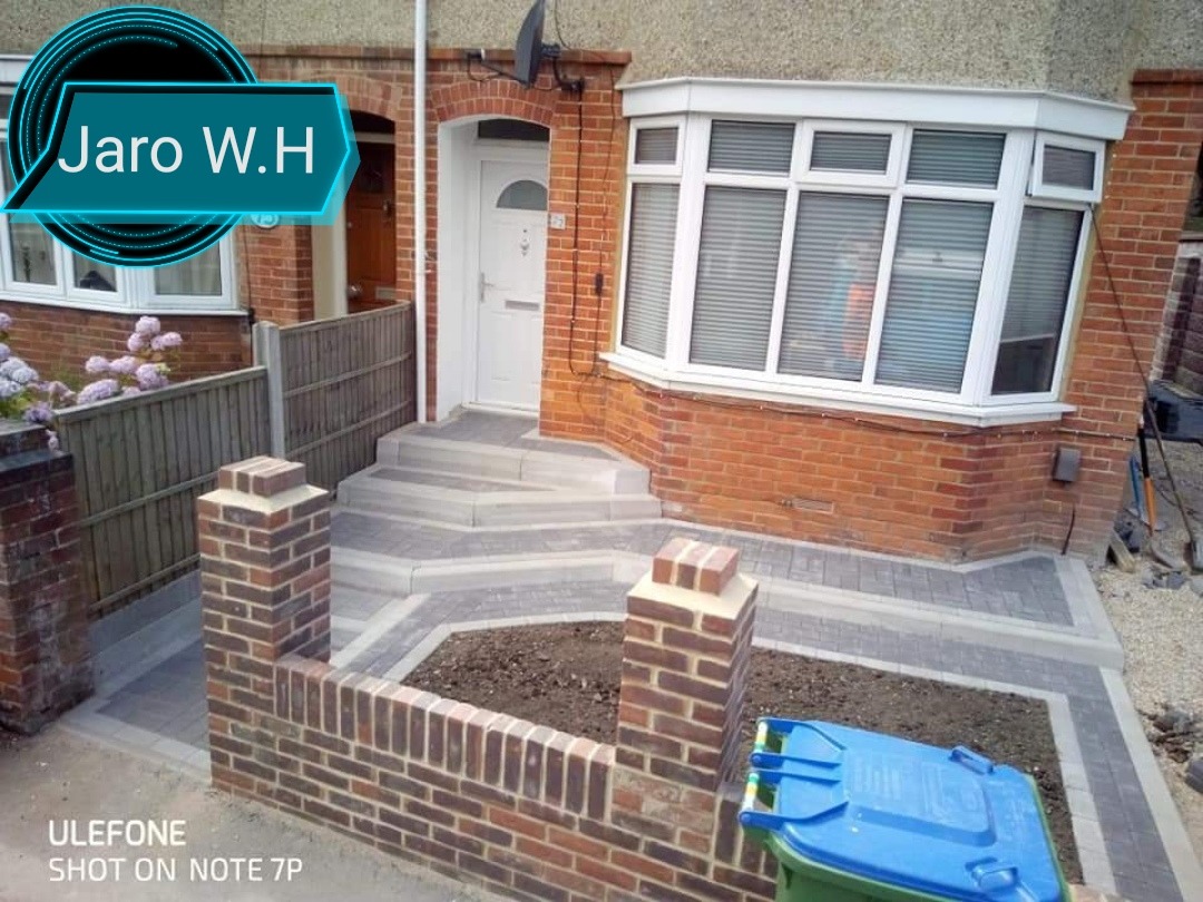 Ułożona kostka brukowa tworząca ścieżkę i schody przed domem z cegły, z widocznym fragmentem trawnika i niebieskim pojemnikiem na śmieci w rogu.