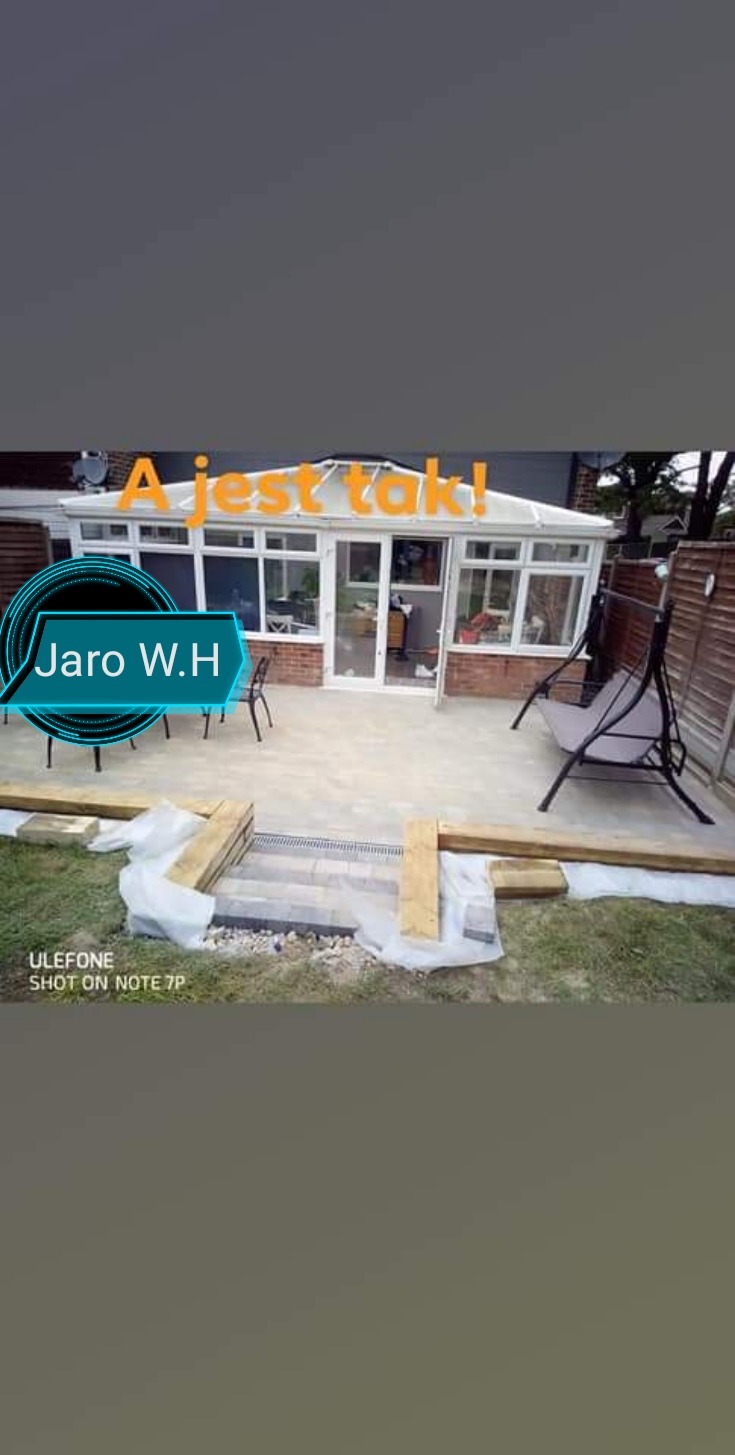 Wykończony taras z jasnych płyt, z odpływem liniowym i schodkami. W tle oranżeria z ceglaną podstawą i huśtawka ogrodowa.