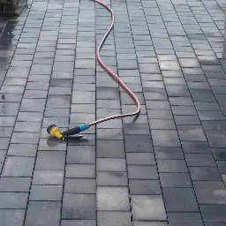 Świeżo ułożona, mokra, szara kostka brukowa w prostokątnym wzorze, z widoczną ubijarką wibracyjną i wężem ogrodowym z żółtą końcówką.