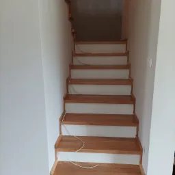 Jasne, drewniane schody z widocznym usłojeniem, zamontowane w białym pomieszczeniu. Częściowo widoczne stopnie wiszące. Kabel elektryczny zwisający pomiędzy stopniami.