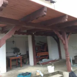 Drewniana konstrukcja z brązowymi, profilowanymi słupami, prawdopodobnie zadaszenie tarasu, w tle widoczne meble ogrodowe i elementy wyposażenia.