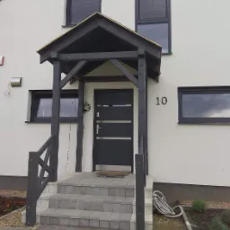 Szare drzwi wejściowe z trzema poziomymi przeszkleniami, zadaszeniem i schodami z szarej kostki, numer domu '10' na białej elewacji.