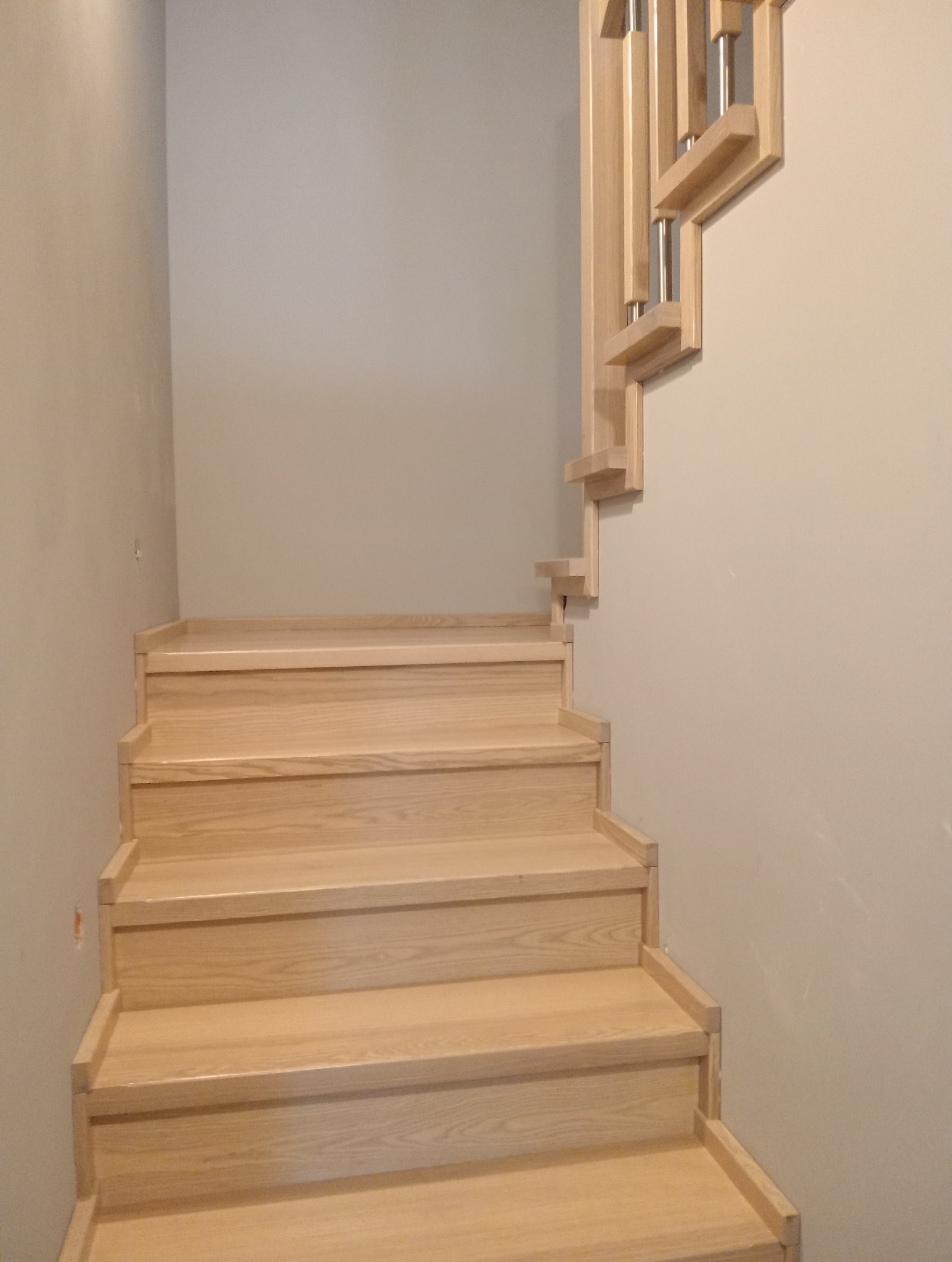 Jasne, drewniane schody z nowoczesną balustradą z elementami metalowymi, widoczne z dołu, przy neutralnej ścianie.