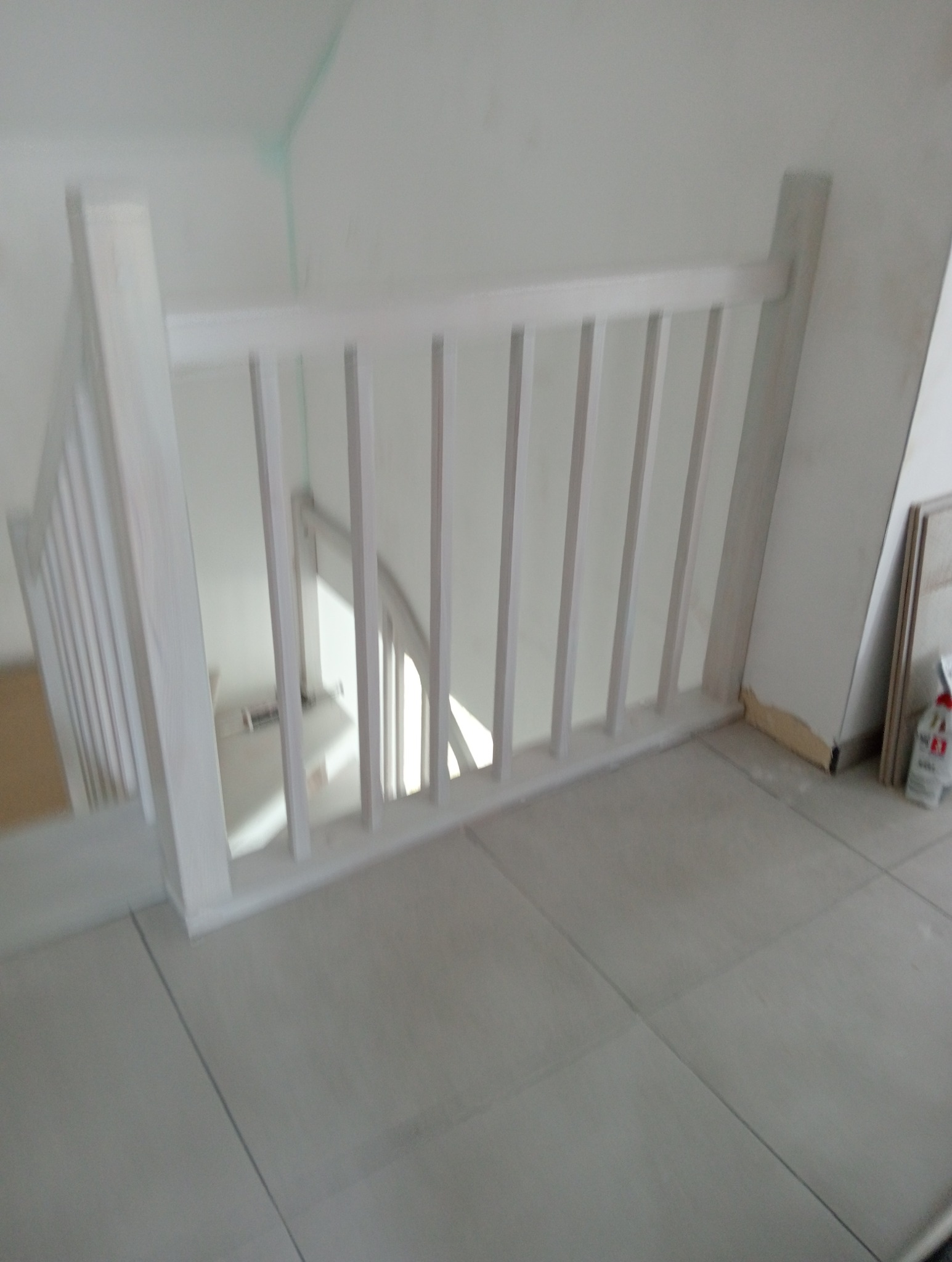 Fragment białej balustrady schodowej z widokiem na schody w dół, na podłodze płytki ceramiczne, widoczne ślady prac wykończeniowych.