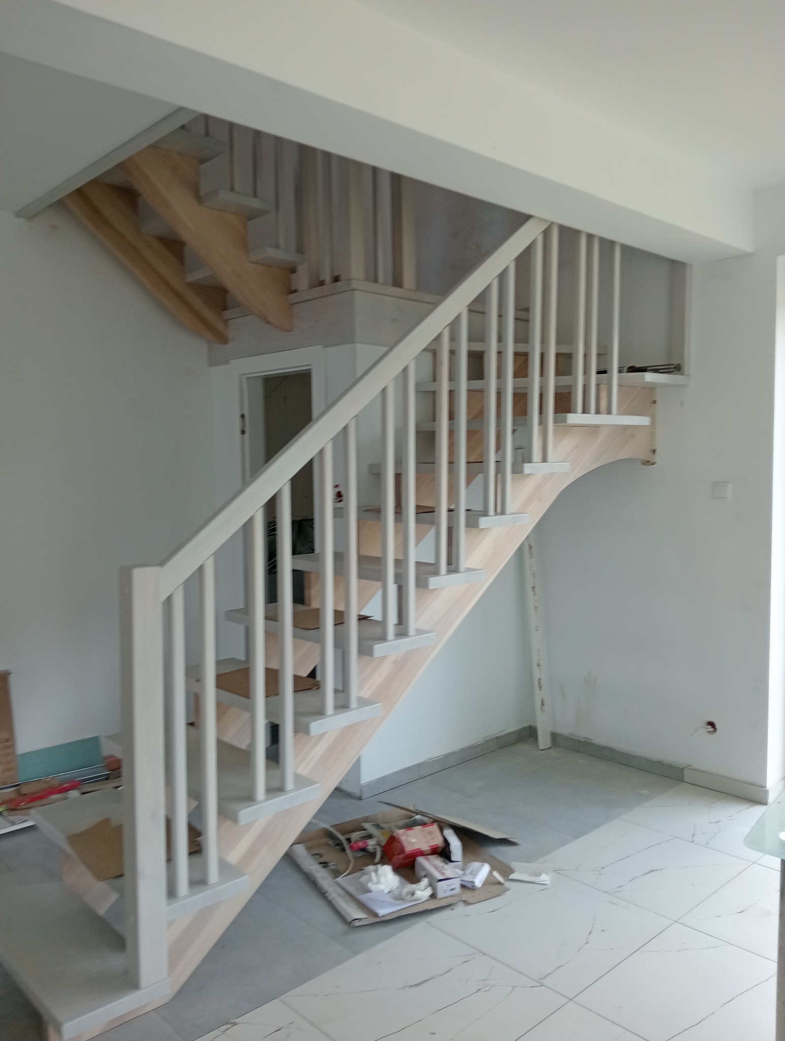 Jasne, drewniane schody z białymi barierkami w trakcie montażu, widoczne narzędzia i materiały budowlane na podłodze.