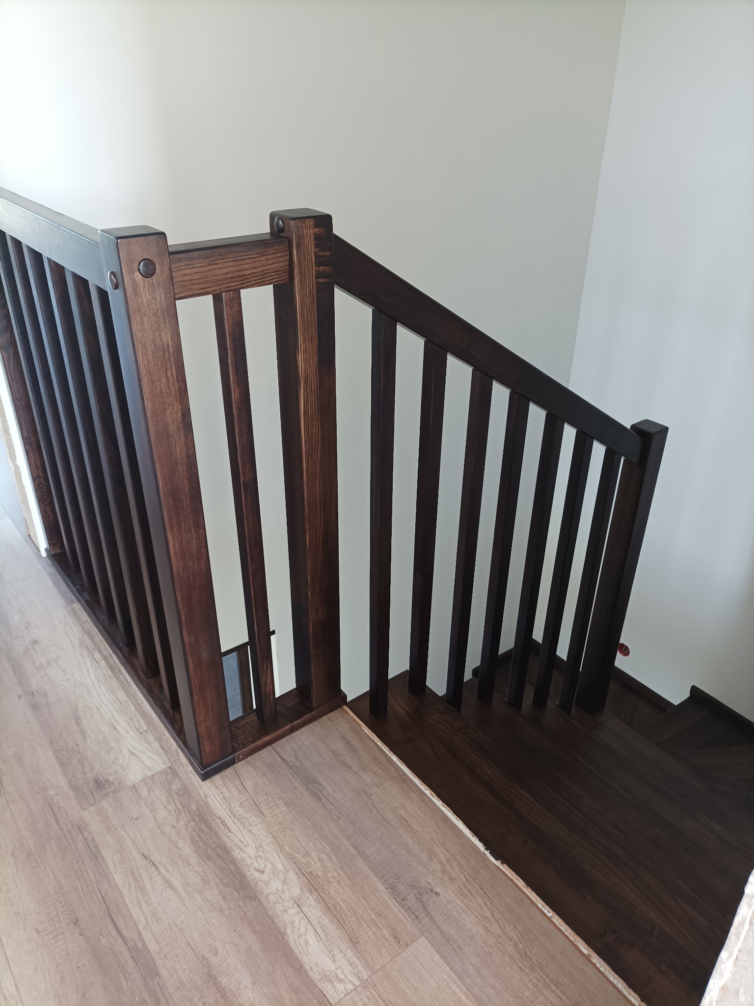 Ciemne, drewniane schody z balustradą w nowoczesnym domu, widok z góry na pierwszy bieg schodów, z parkietem na podłodze i białymi ścianami.