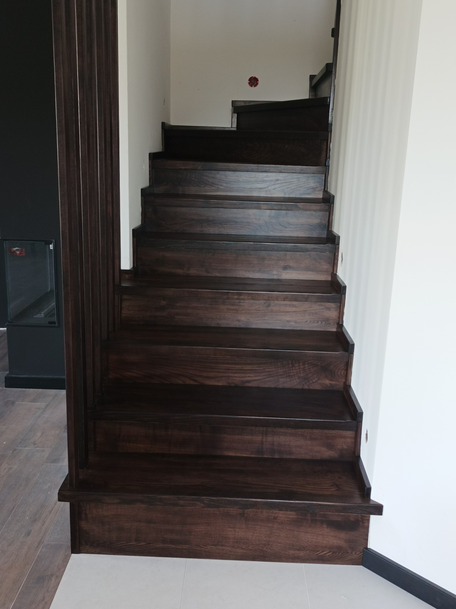 Ciemne, drewniane schody z widoczną strukturą drewna, w nowoczesnym wnętrzu z jasnymi ścianami i ciemnymi elementami dekoracyjnymi.