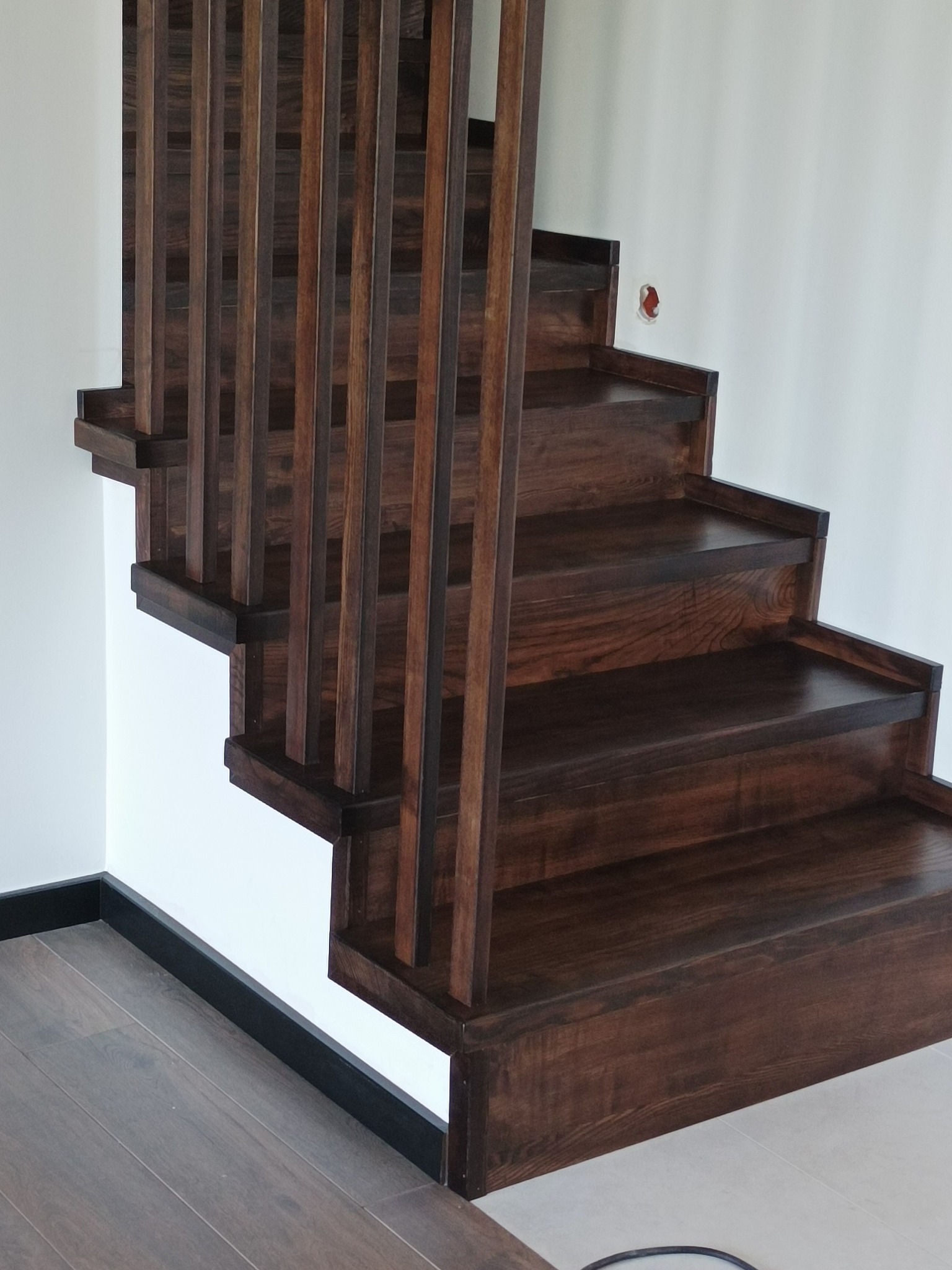 Ciemnobrązowe schody z drewnianymi, pionowymi balustradami, widoczne od dołu, z fragmentem podłogi i białej ściany z niedokończonym gniazdkiem elektrycznym.