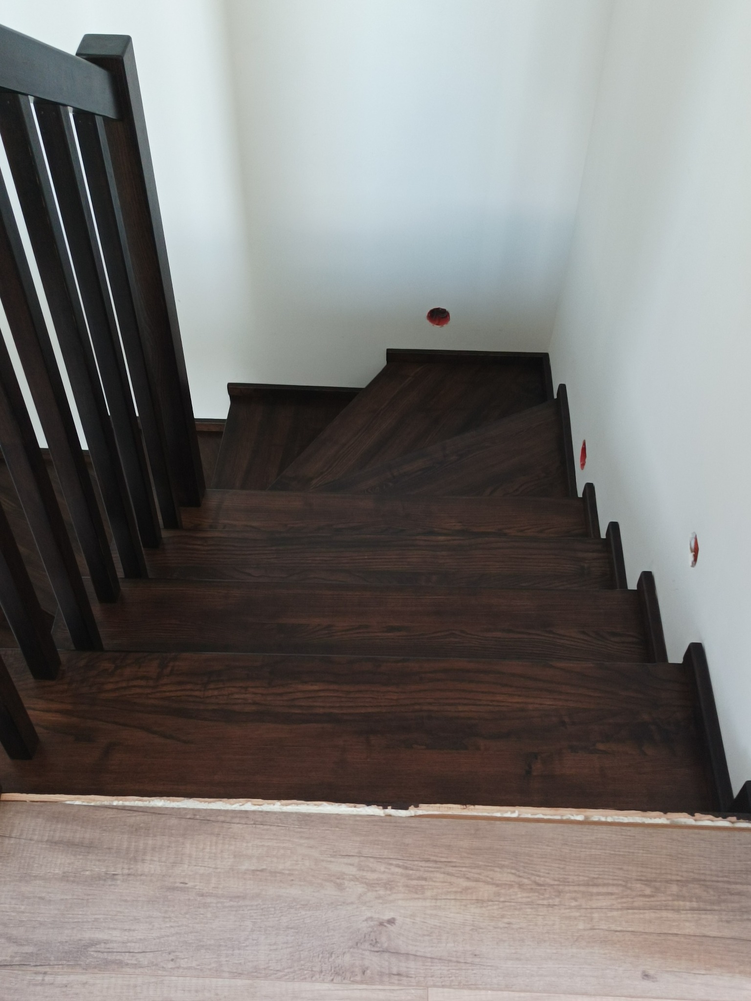 Widok z góry na ciemne, drewniane schody z minimalistyczną balustradą, z widocznymi otworami na gniazdka elektryczne w ścianie i pianką montażową u podstawy schodów.