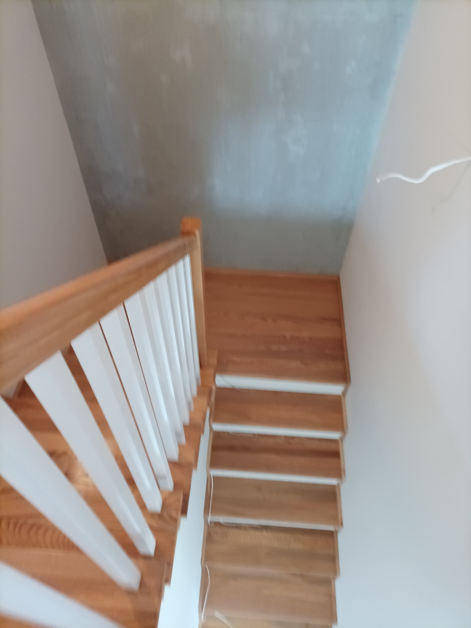 Drewniane schody z jasnymi, białymi balustradami w trakcie montażu, surowy betonowy sufit w tle, widok z góry.