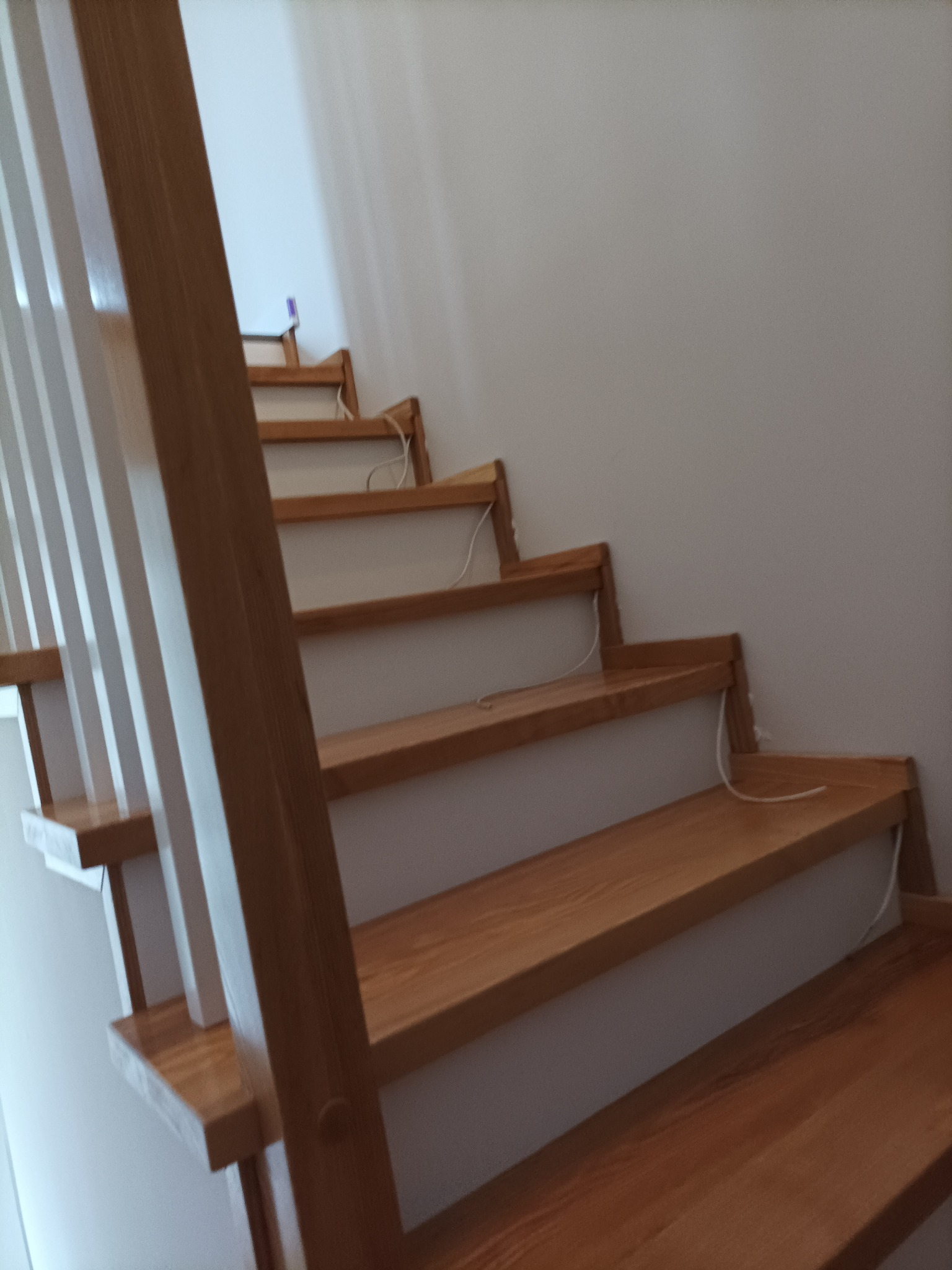 Nowoczesne schody drewniane z białymi podstopnicami i jasnymi balustradami, widoczne z dołu, z przewodami wystającymi spod stopni.