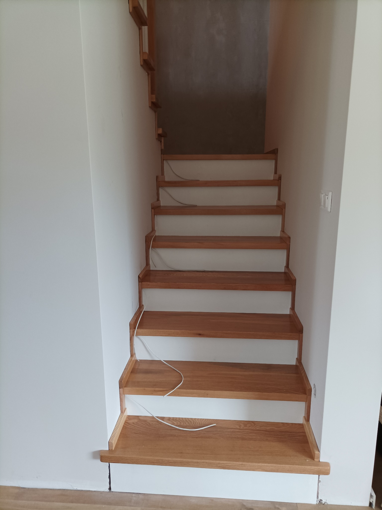 Jasne, drewniane schody z widocznym usłojeniem, zamontowane w białym pomieszczeniu. Częściowo widoczne stopnie wiszące. Kabel elektryczny zwisający pomiędzy stopniami.
