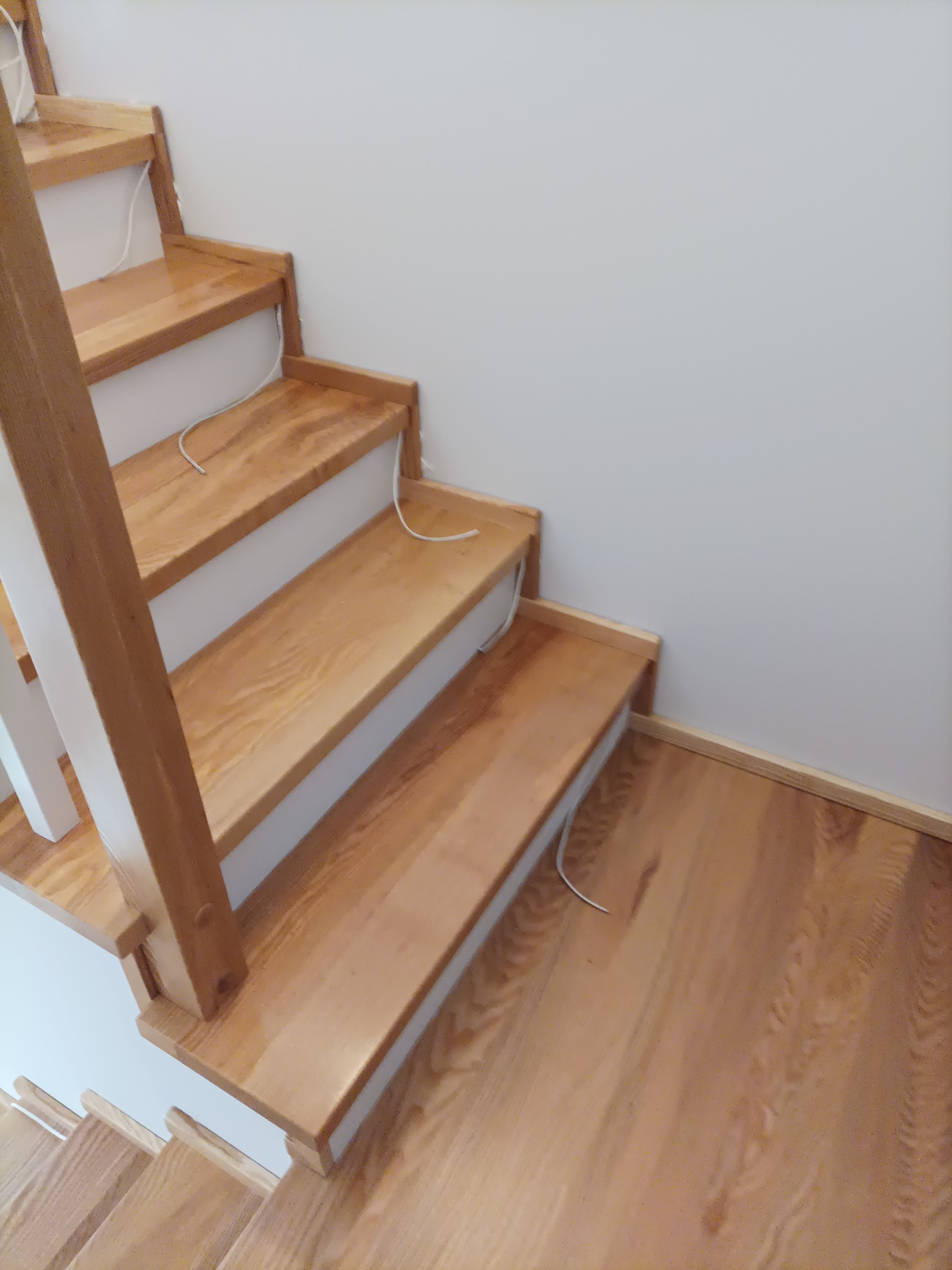 Jasne, drewniane schody z białymi podstopnicami i balustradą, widok z góry na dół, z widocznymi przewodami elektrycznymi.