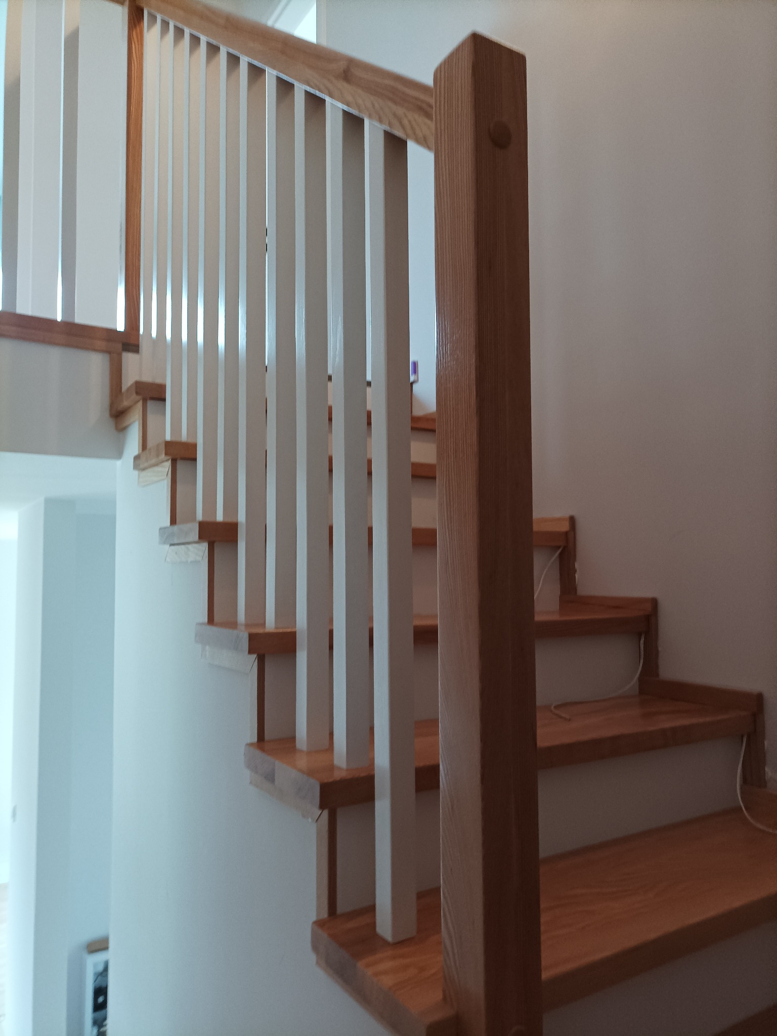 Jasne schody z drewnianymi stopniami i białymi balustradami, widok z dołu, częściowo widoczne kable pod stopniami.
