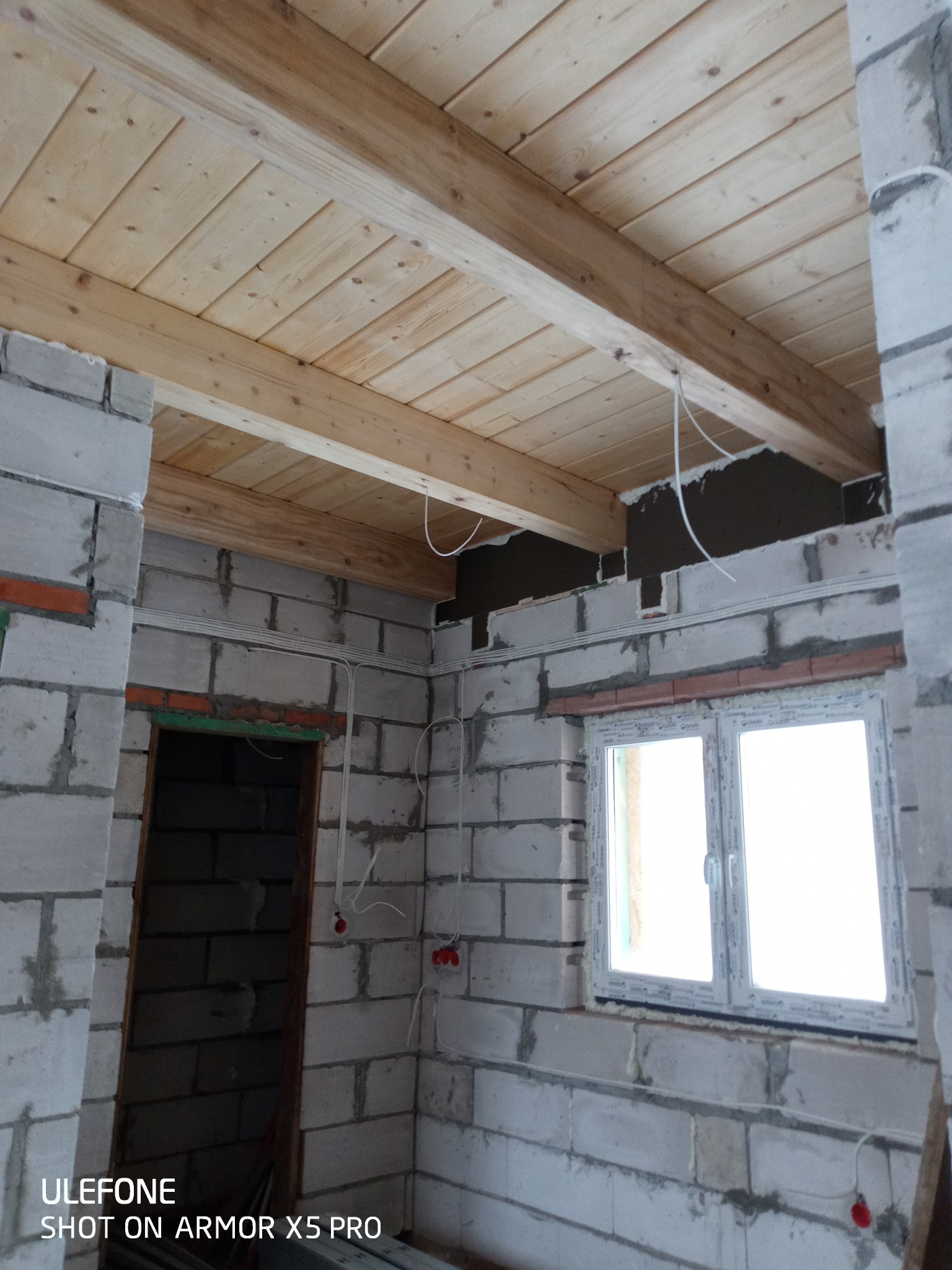 Surowy stan wnętrza w trakcie prac: ściany z betonu komórkowego, drewniany sufit podwieszany, widoczne przewody elektryczne i puszki, okno PCV.