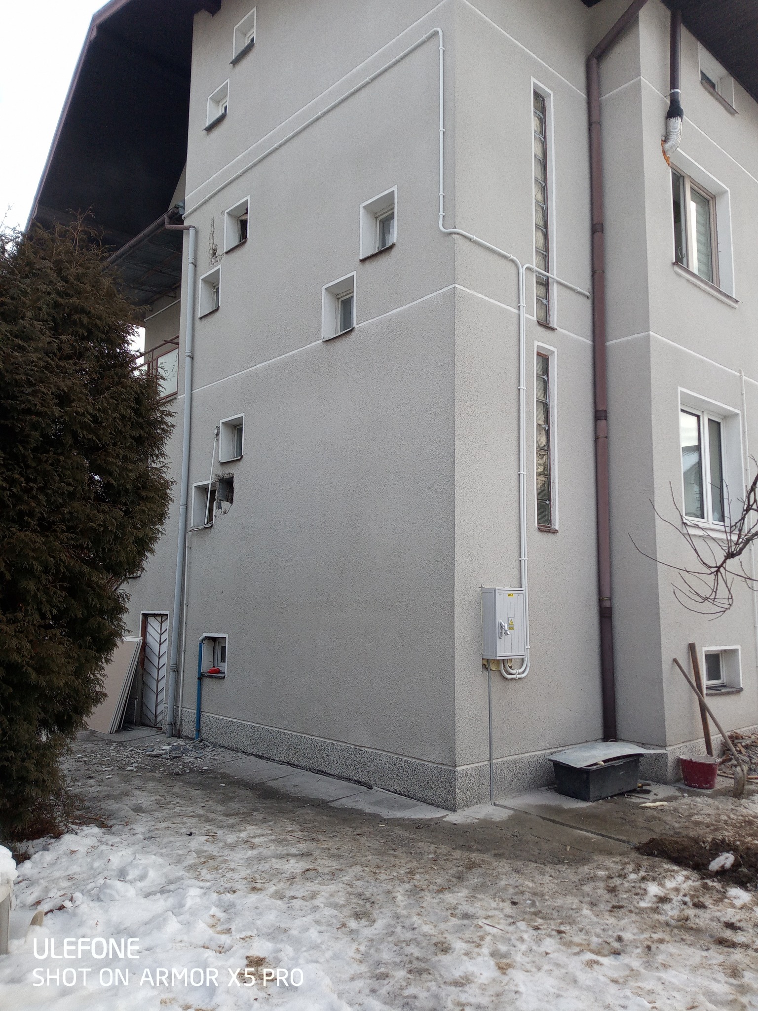 Szary budynek z widoczną skrzynką elektryczną na elewacji, rurami prowadzonymi po ścianie, uszkodzonym oknem i śladami prac remontowych na zewnątrz.