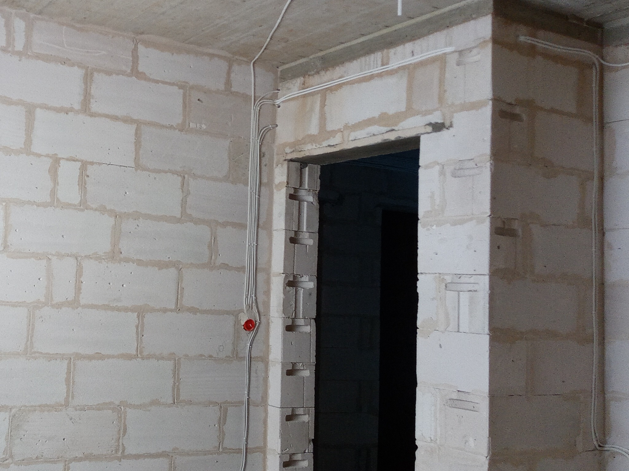 Wewnętrzne ściany z betonu komórkowego w trakcie prac instalacyjnych, widoczne przewody elektryczne prowadzone natynkowo, czerwona puszka instalacyjna i surowy otwór drzwiowy.