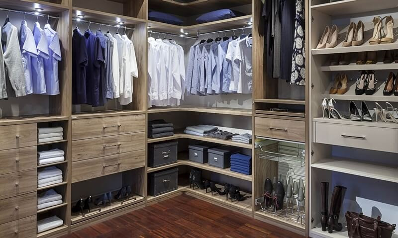 Przestronna szafa wnękowa z drewna w odcieniach beżu, z oświetleniem LED, wyposażona w półki z ubraniami, szuflady, pudełka i wysuwany kosz na buty, prezentująca uporządkowaną garderobę.