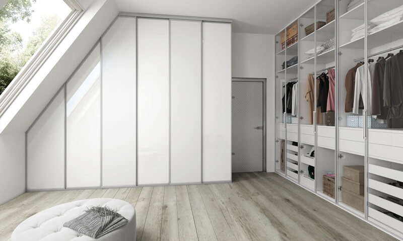 Biała, nowoczesna garderoba w skosie poddasza z przesuwnymi drzwiami, uzupełniona o pufę z narzutą i drewnianą podłogę, oświetlona przez okno dachowe.