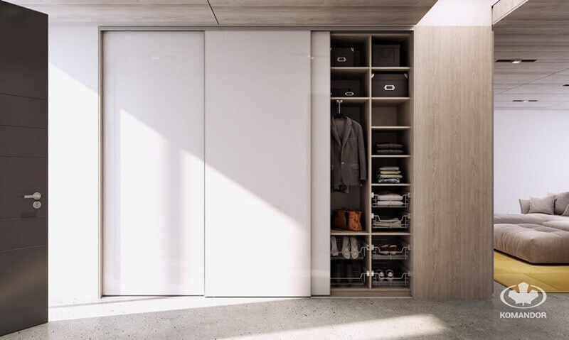 Nowoczesna szafa z przesuwnymi, białymi drzwiami, widoczne półki z ubraniami i akcesoriami, oświetlenie punktowe, podłoga betonowa, w tle część sofy i logo firmy Komandor.