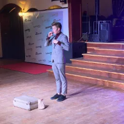 Młody mężczyzna w garniturze na scenie, w tle baner reklamowy Studniówka 2020, obok niego walizka i papierowa torba, prawdopodobnie podczas występu.