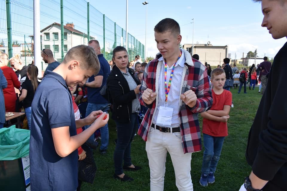 Młody mężczyzna w kraciastej koszuli prezentuje trik z małym czerwonym przedmiotem, otoczony przez publiczność na tle zielonego trawnika.