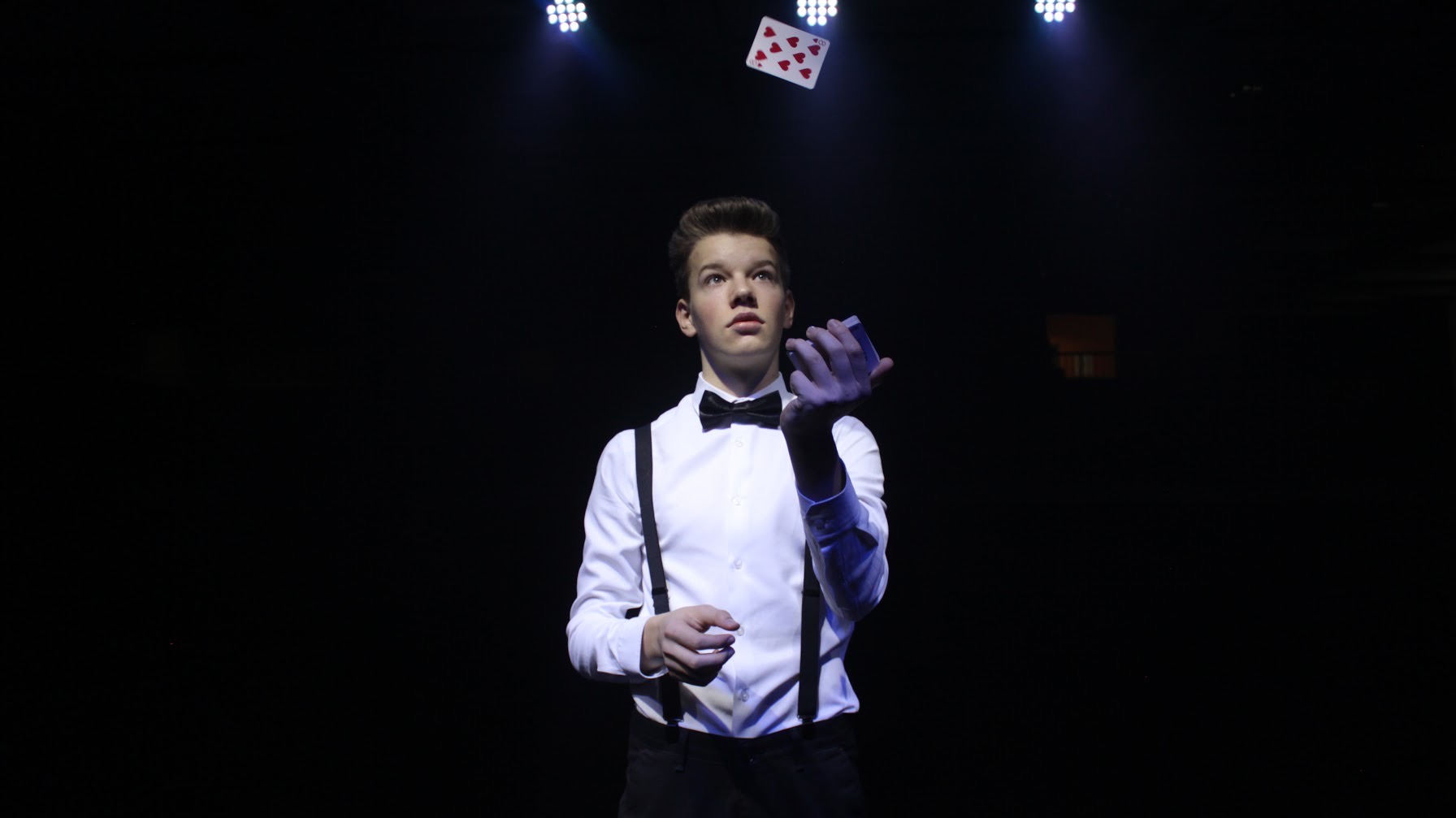 Młody iluzjonista w białej koszuli z muchą i szelkami, karta do gry unosi się nad jego ręką, oświetlenie sceniczne w tle.