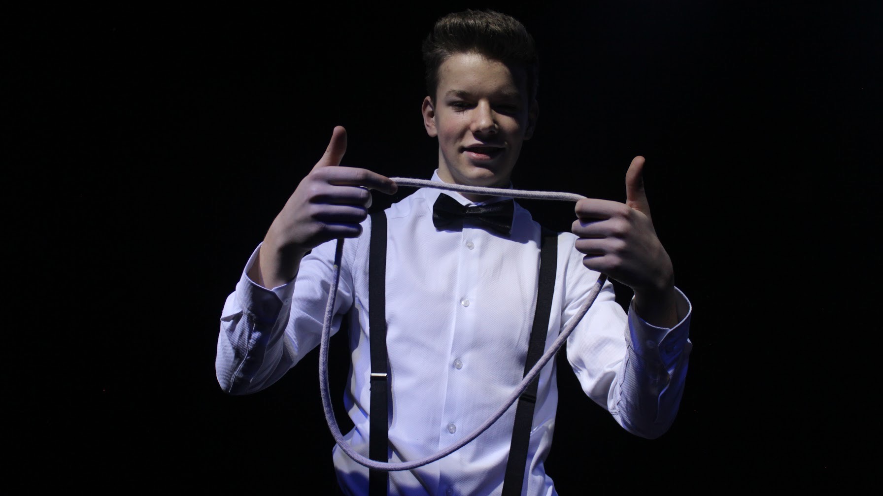 Młody iluzjonista w białej koszuli z muszką i szelkami, prezentuje trik z liną na czarnym tle, trzymając kciuki w górę.