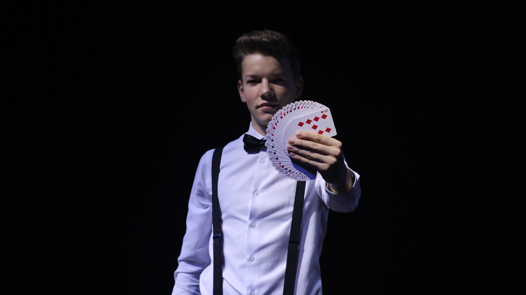 Młody mężczyzna w białej koszuli, muszce i szelkach prezentuje wachlarz kart do gry na czarnym tle.