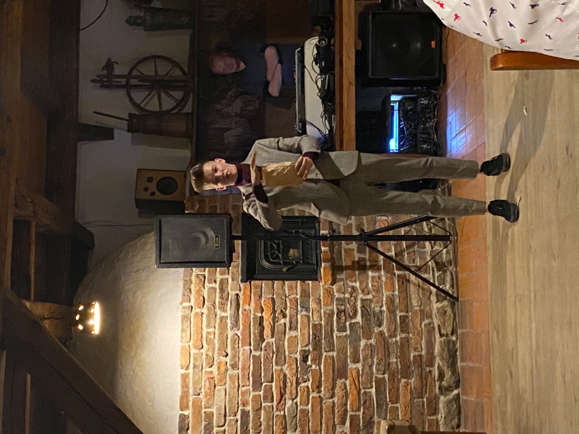 Młody mężczyzna w garniturze, stojący przed ceglaną ścianą z głośnikiem, trzymający brązową papierową torbę, prawdopodobnie podczas występu.