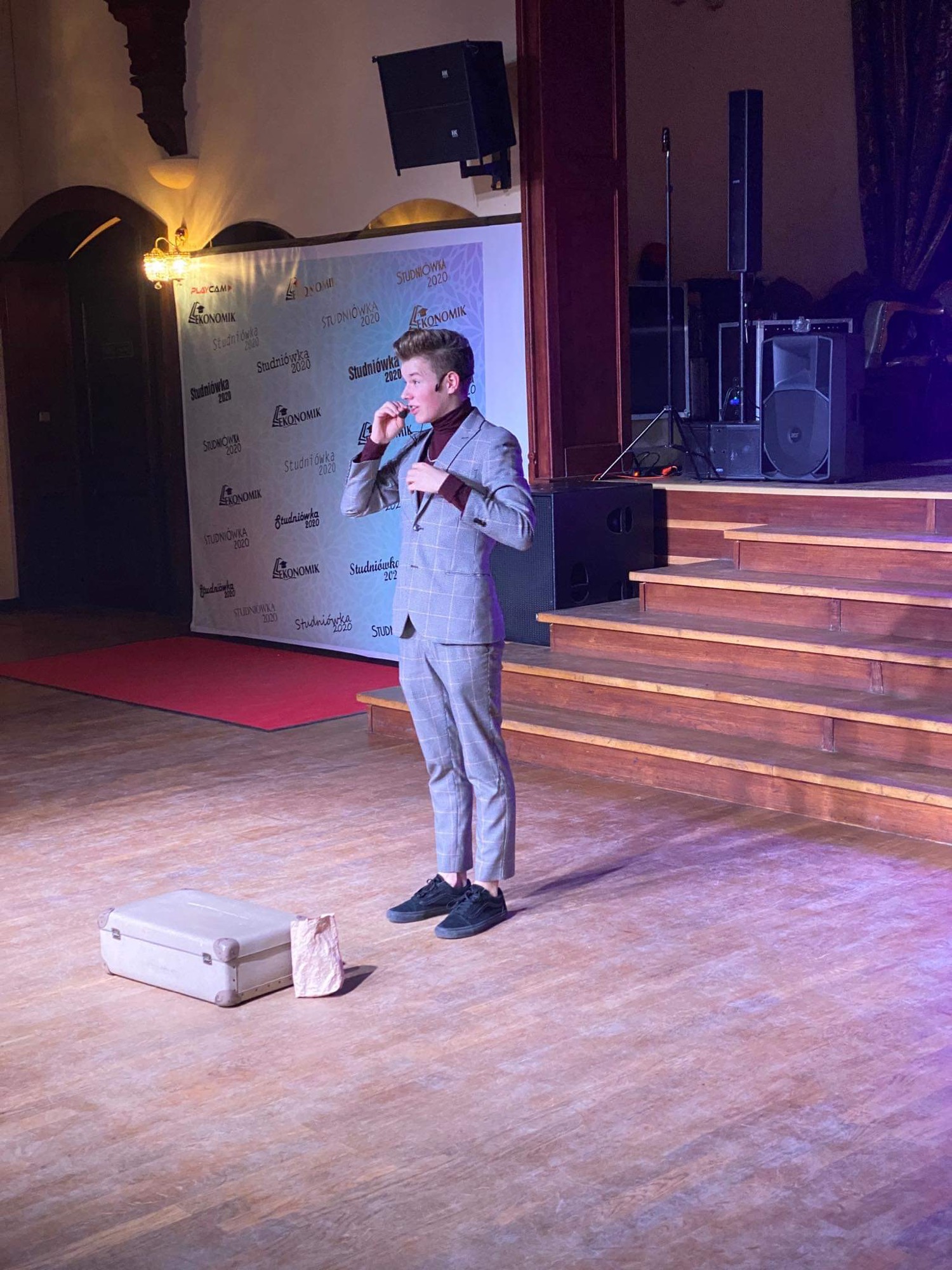 Młody mężczyzna w garniturze na scenie, w tle baner reklamowy Studniówka 2020, obok niego walizka i papierowa torba, prawdopodobnie podczas występu.