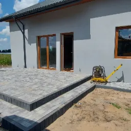 Nowoczesny taras z szarej kostki brukowej z trzema stopniami, przylegający do szarego domu z drewnianymi oknami, ubijarka wibracyjna obok, widok na pole w tle.