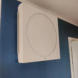 Biała, kwadratowa jednostka centralna z okrągłym panelem na niebieskiej ścianie, widoczne logo LG, obok białe drzwi.