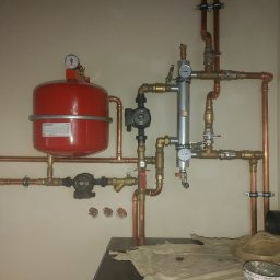 Instalacje Sanitarne Michał Szczepaniak - Nowa instalacja grzewcza z czerwonym zbiornikiem wyrównawczym, miedzianymi rurami i manometrami, widoczne ślady montażu i resztki pakuł na podłodze.
