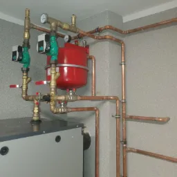 Szara kotłownia z piecem, czerwonym zbiornikiem wyrównawczym i miedzianymi rurami, widoczne manometry i pompy obiegowe z zielonymi obudowami.