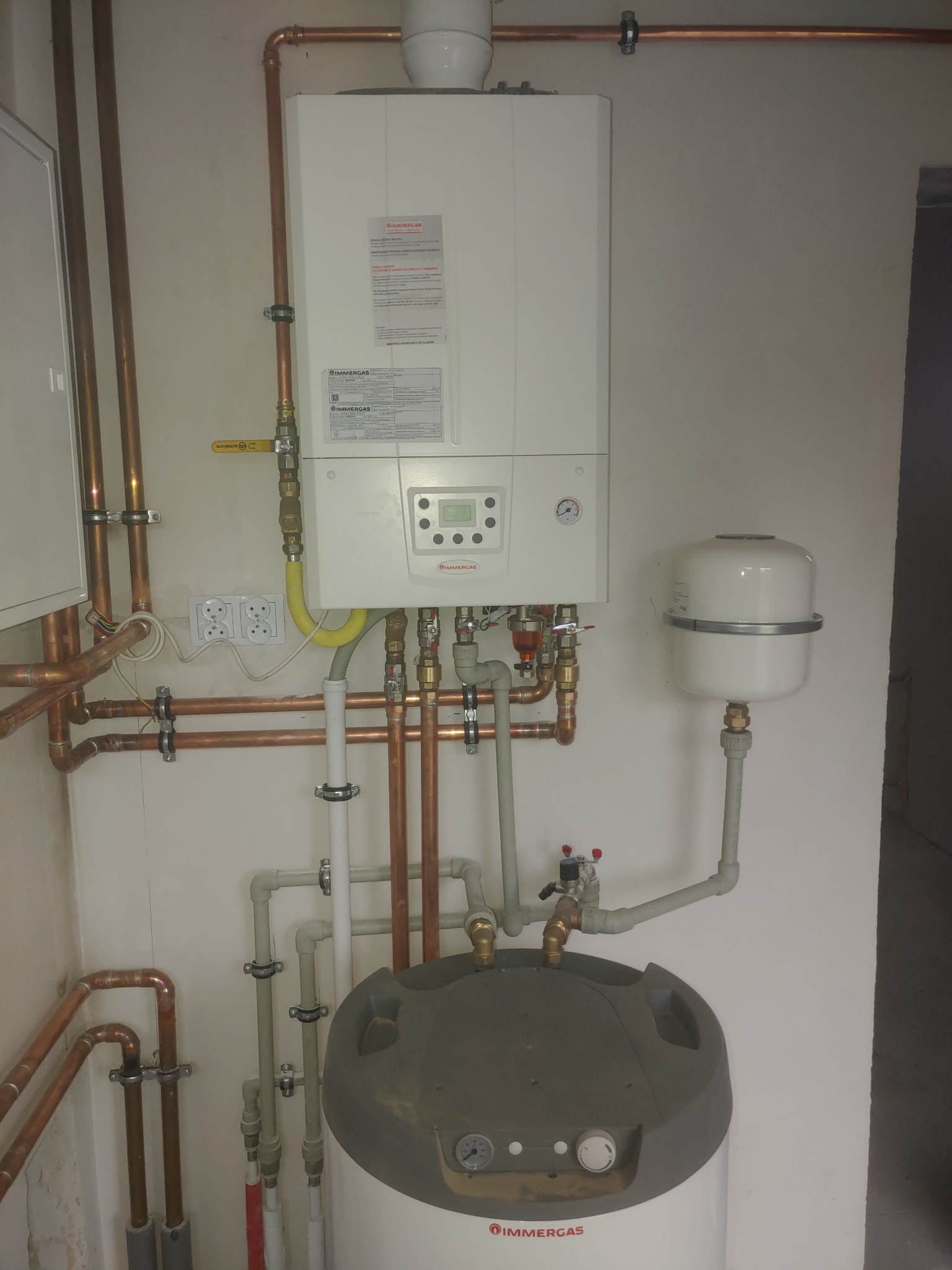 Biały, wiszący piec gazowy Immergas z miedzianymi rurami, podłączony do szarego zbiornika, widoczne zawory i manometr.