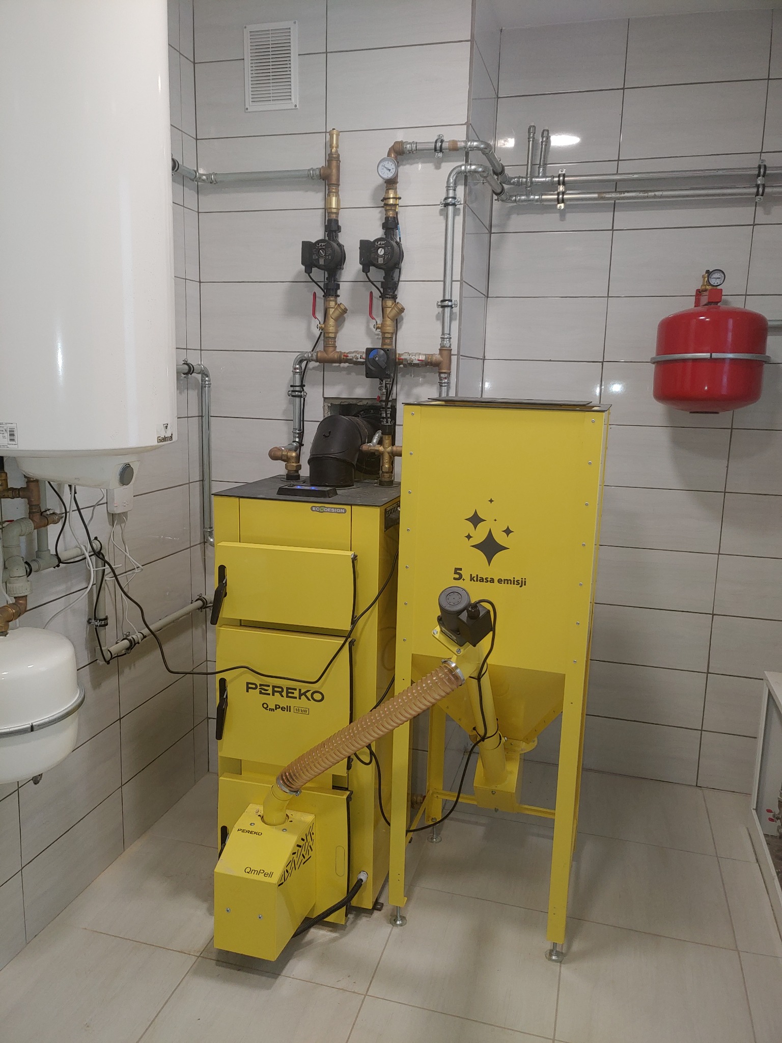 Żółty piec na pellet Pereko QmPell 16kW z podajnikiem, instalacja CO z białym zasobnikiem i czerwonym naczyniem wzbiorczym w kotłowni wyłożonej białymi płytkami.