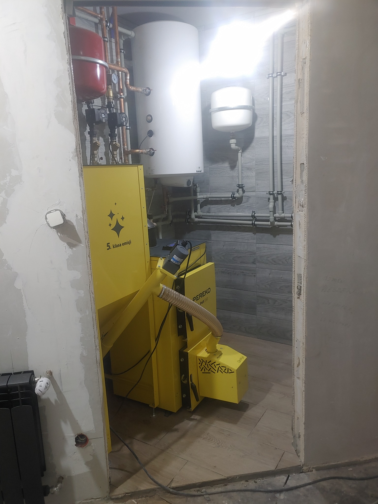 Żółty piec na pellet marki PEREKO w trakcie instalacji, widoczne rury instalacji grzewczej, zbiornik buforowy i częściowo wykończona ściana z instalacją elektryczną.