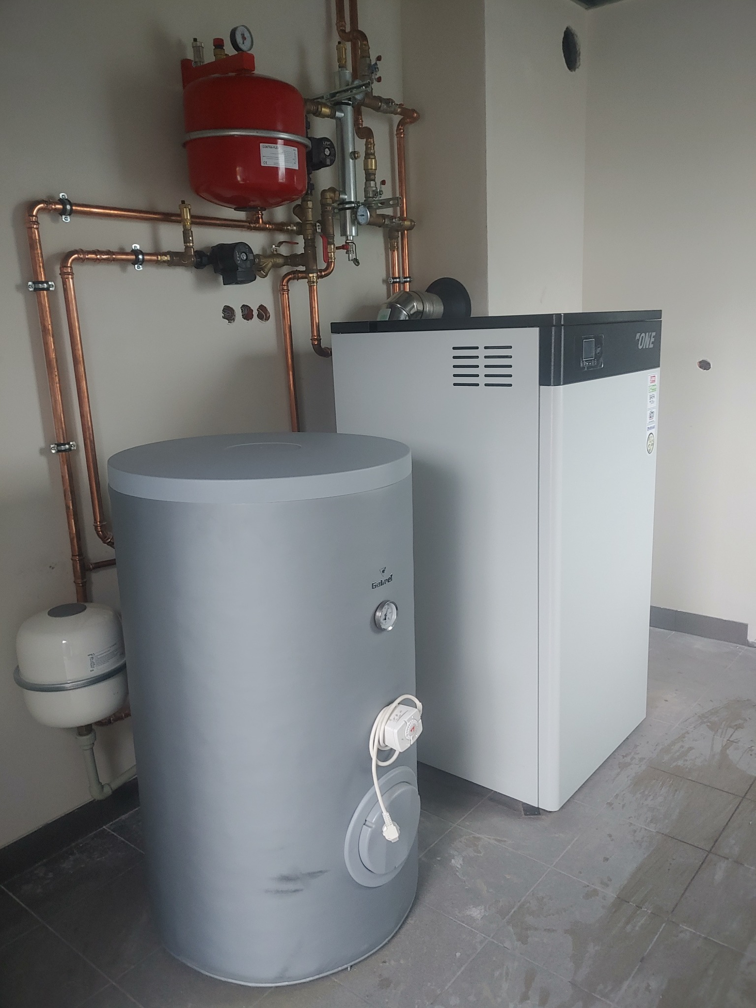 Nowoczesna kotłownia z szarym zasobnikiem ciepłej wody, białym piecem z czarnym panelem sterowania i miedzianą instalacją, widoczne ślady po wierceniu w ścianie.