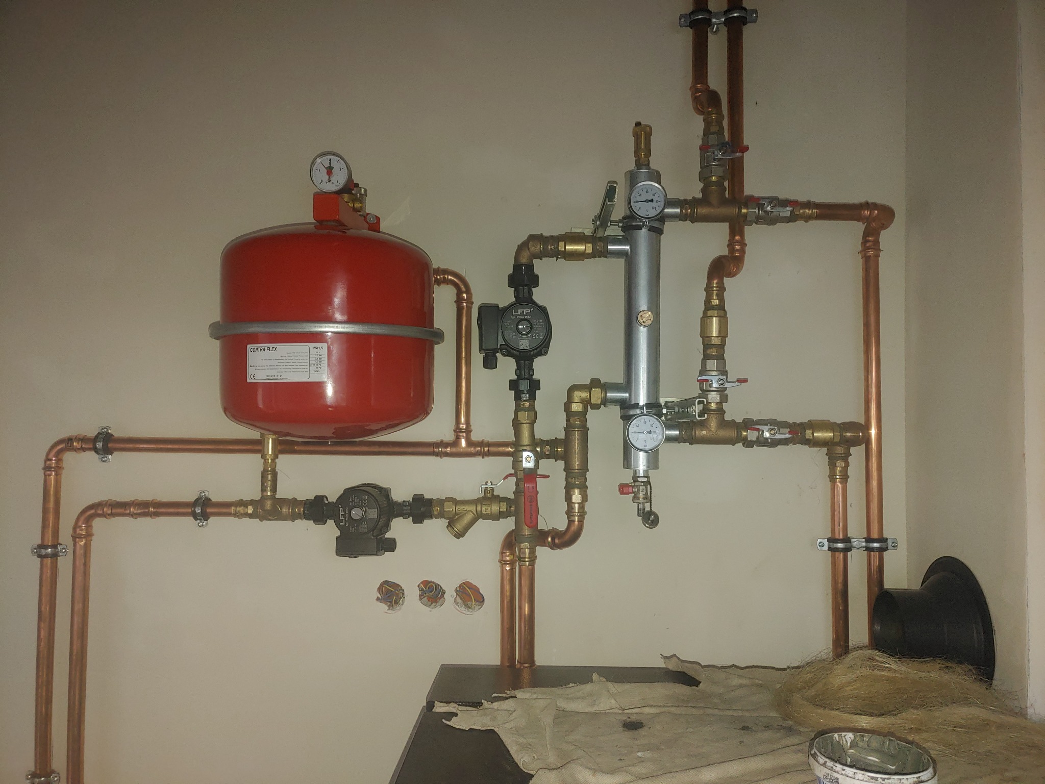Nowa instalacja grzewcza z czerwonym zbiornikiem wyrównawczym, miedzianymi rurami i manometrami, widoczne ślady montażu i resztki pakuł na podłodze.