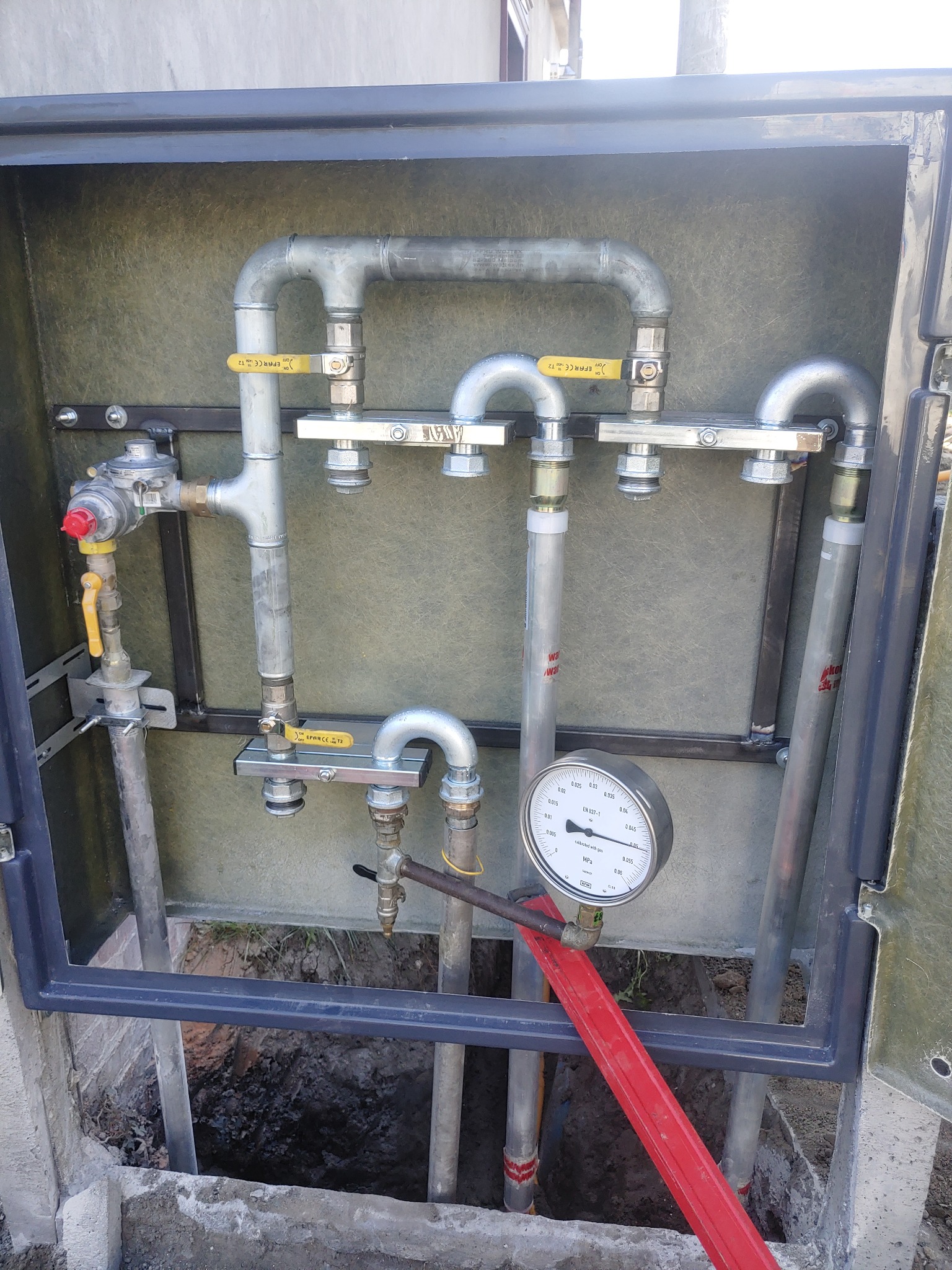 Szafka gazowa z zaworami, manometrem i rurami, widok z przodu, w tle wykop ziemny.