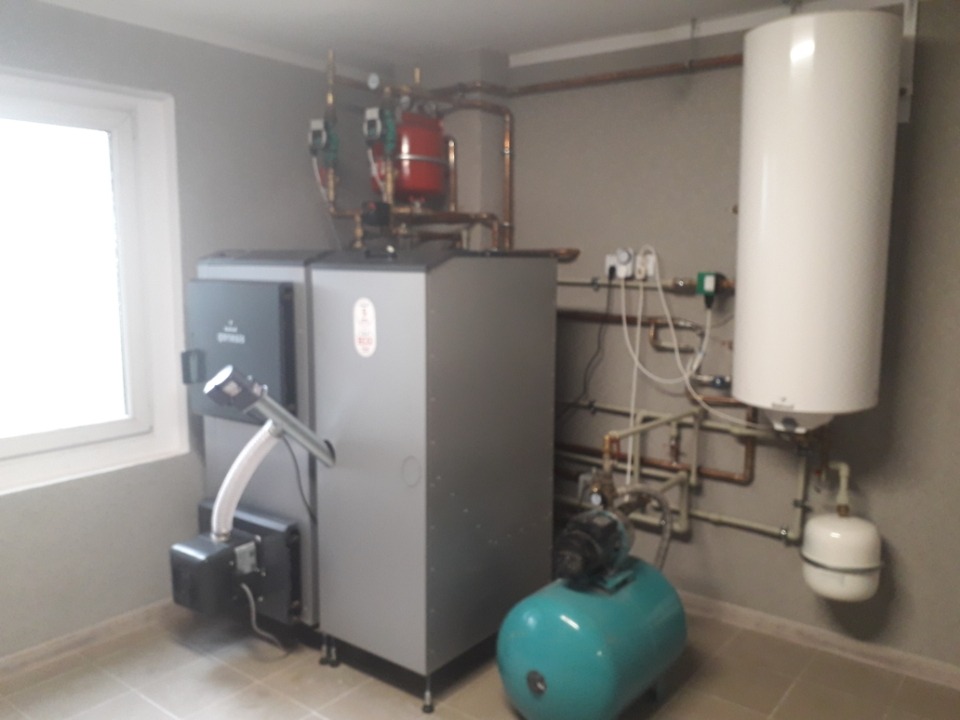 Kompletna kotłownia z kotłem na paliwo stałe, zasobnikiem ciepłej wody i instalacją hydrauliczną w pomieszczeniu z oknem.