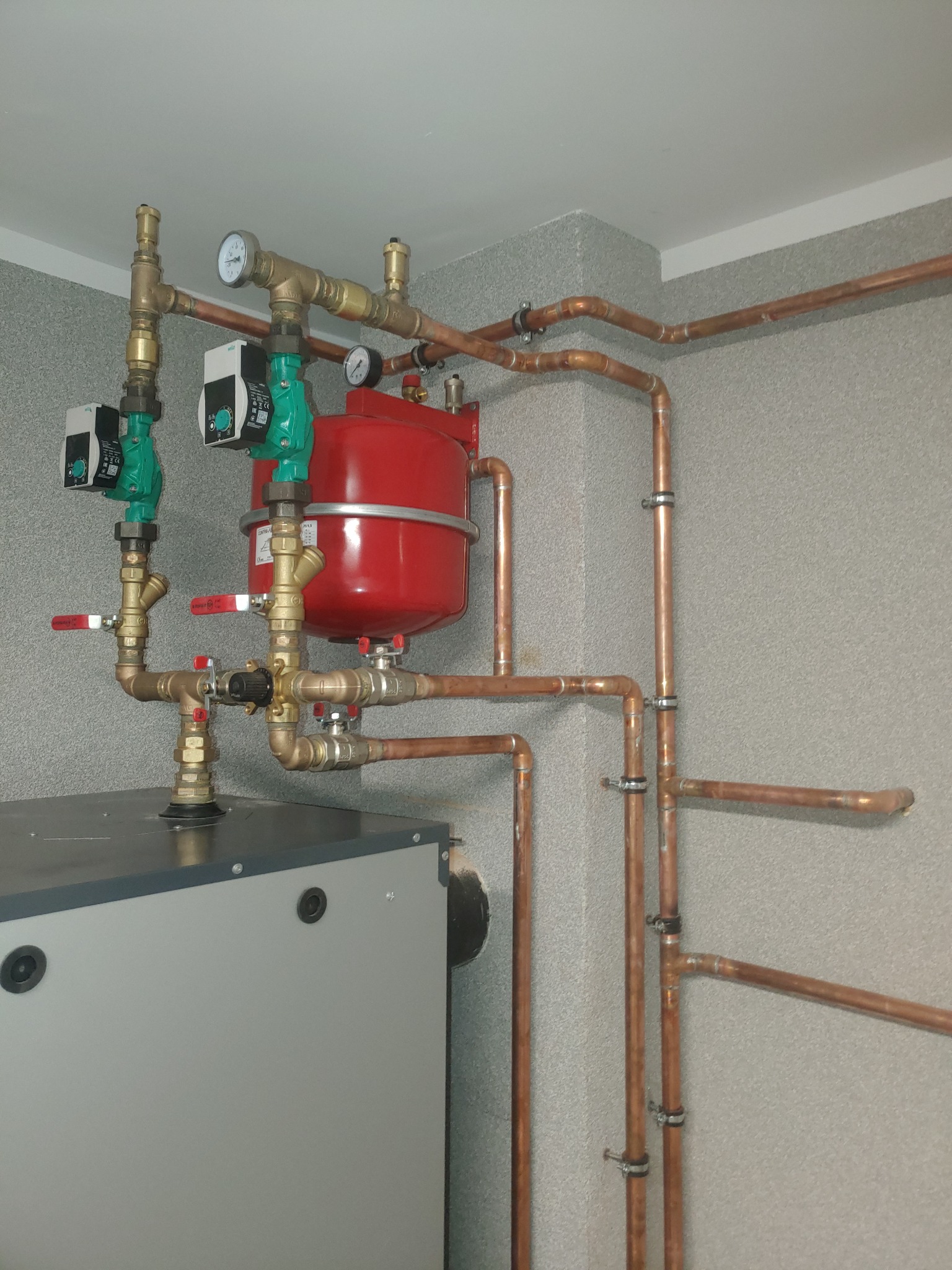 Szara kotłownia z piecem, czerwonym zbiornikiem wyrównawczym i miedzianymi rurami, widoczne manometry i pompy obiegowe z zielonymi obudowami.