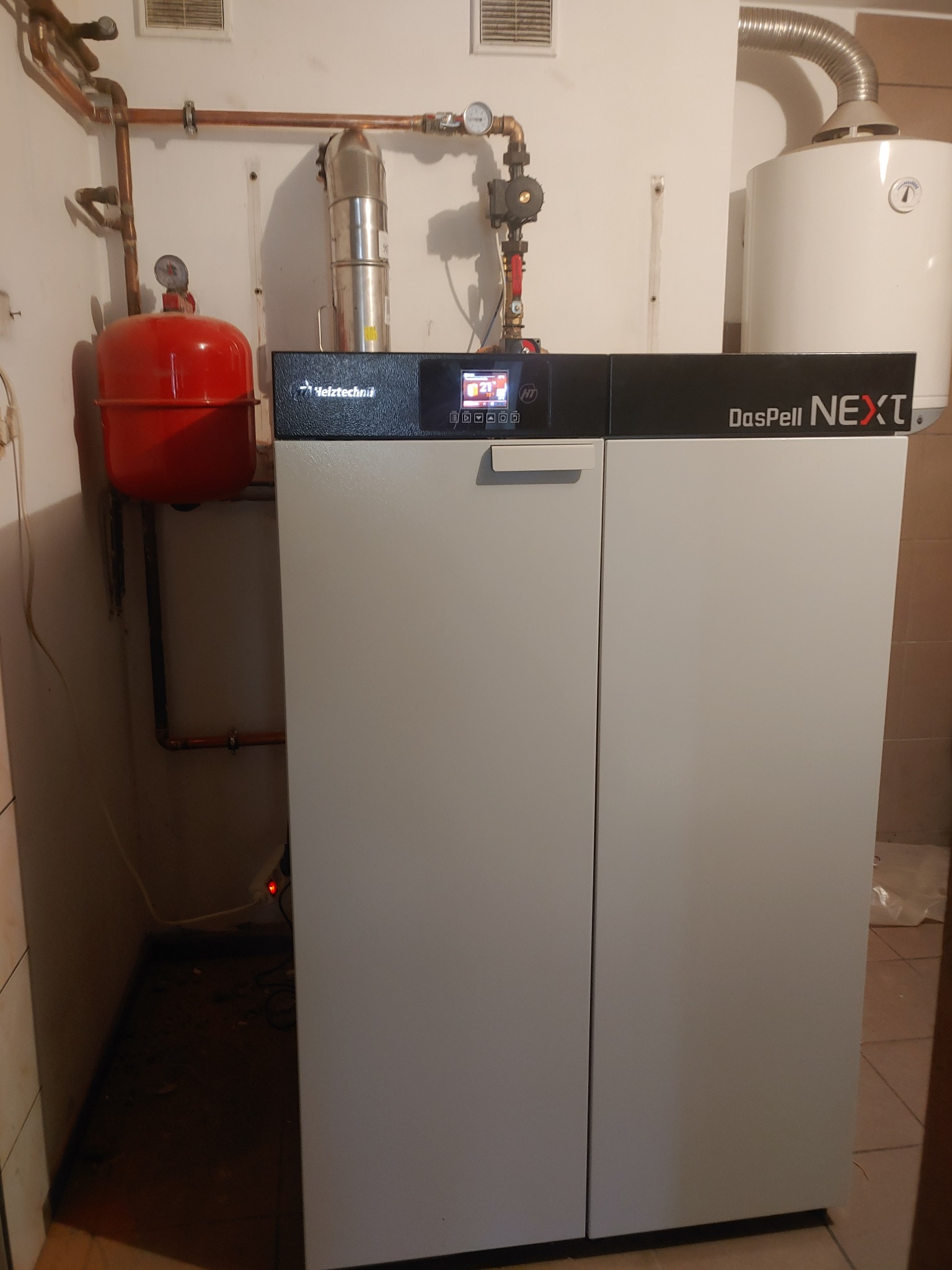 Kompaktowy piec DasPell NEXT z czerwonym zbiornikiem wyrównawczym i instalacją hydrauliczną z miedzianych rur w kotłowni.