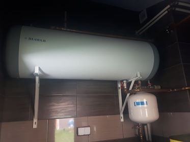 Poziomy, biały bojler marki Biawar zamontowany na ścianie z brązowych płytek, widoczne rury miedziane i mały zbiornik wyrównawczy. Ujęcie z dołu, lekko zniekształcające proporcje.