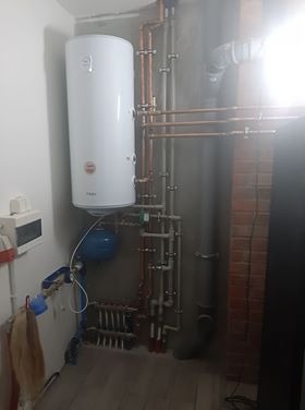 Biały bojler zawieszony na szarej ścianie, połączony miedzianymi rurami z innymi elementami instalacji w pomieszczeniu o surowym wykończeniu. Widoczne liczne zawory i złączki. Część rur biegnie...