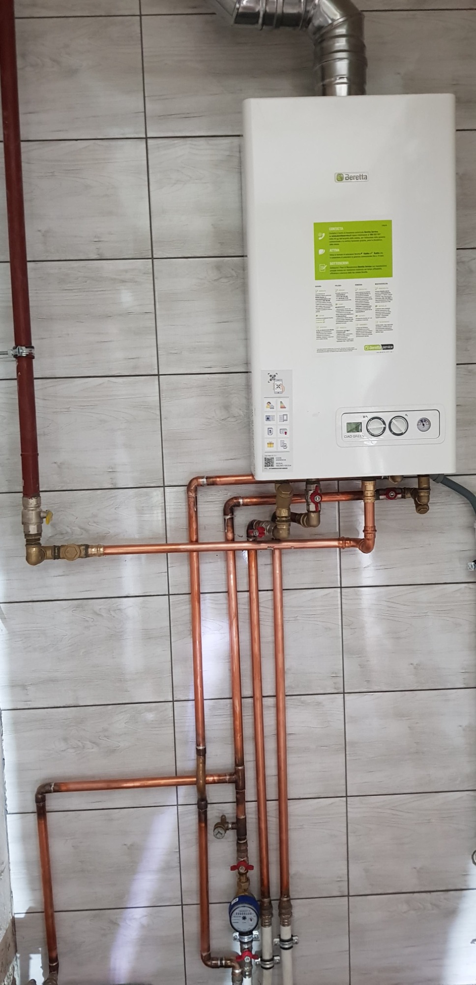 Biały piec gazowy Beretta z podłączonymi miedzianymi rurami, zamontowany na ścianie z szarymi płytkami imitującymi drewno. Widoczny manometr i zawory.